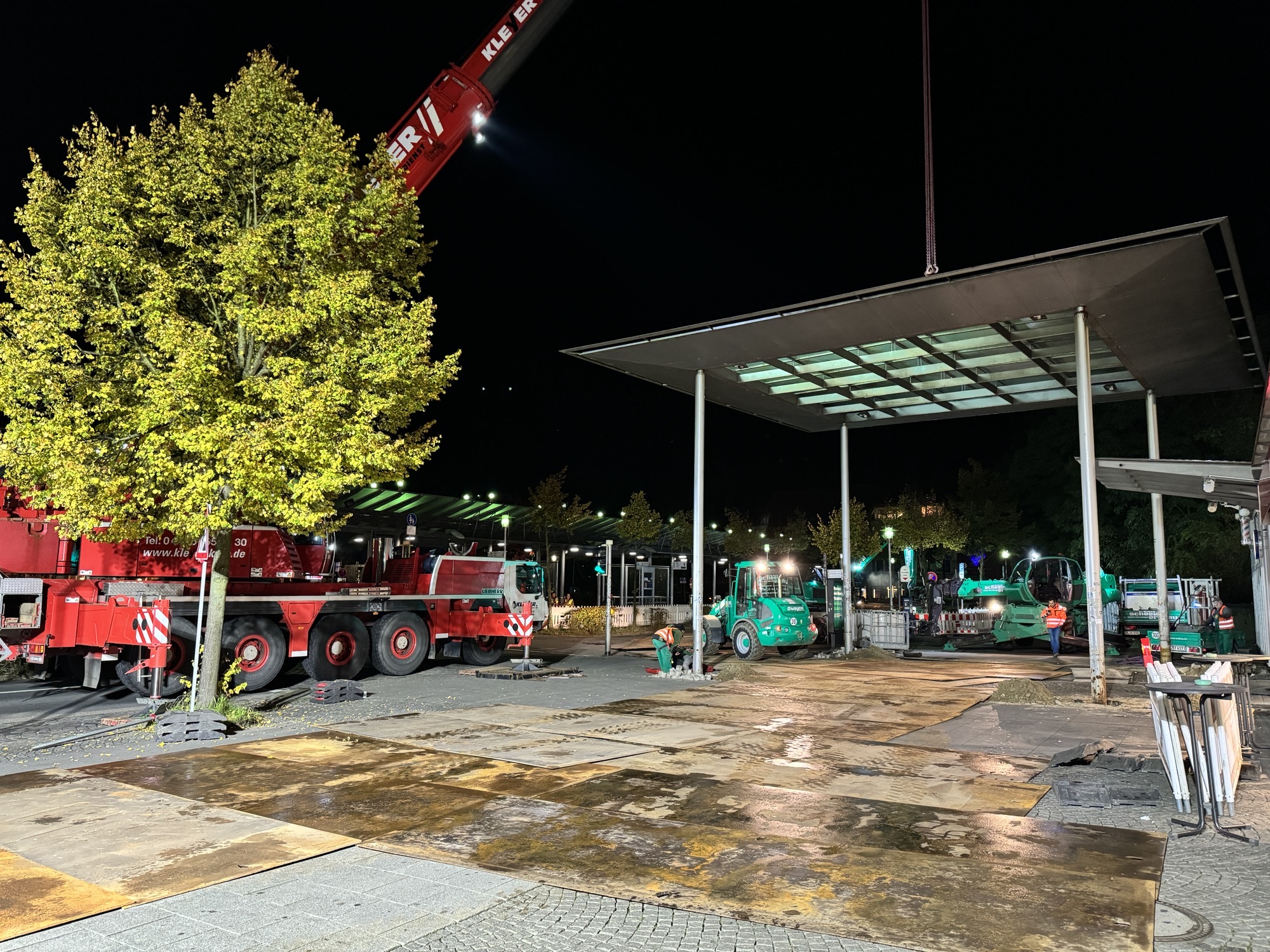 Baustelle bei Nacht mit verschiedenen Fahrzeugen, darunter ein roter Kran und ein grüner Bagger, unter einem beleuchteten Vordach.